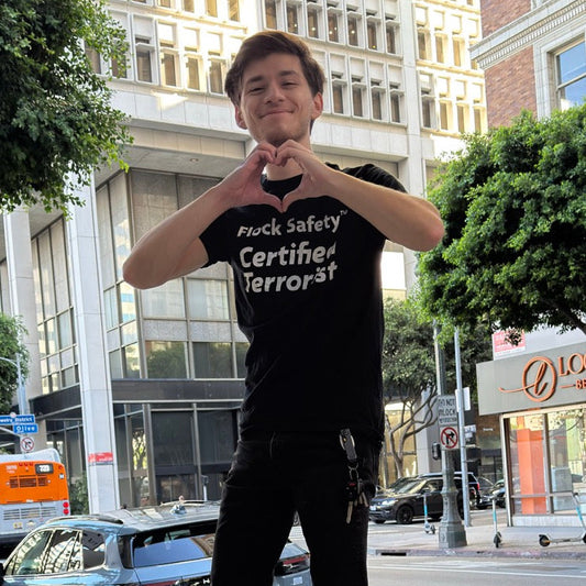 A man with a goofy grin wearing a black t-shirt with 'Flock Safety Certified Terrorist' printed on it stands on a food delivery robot on a city street, making a heart shape with his hands. Cars and city buildings are visible in the background.